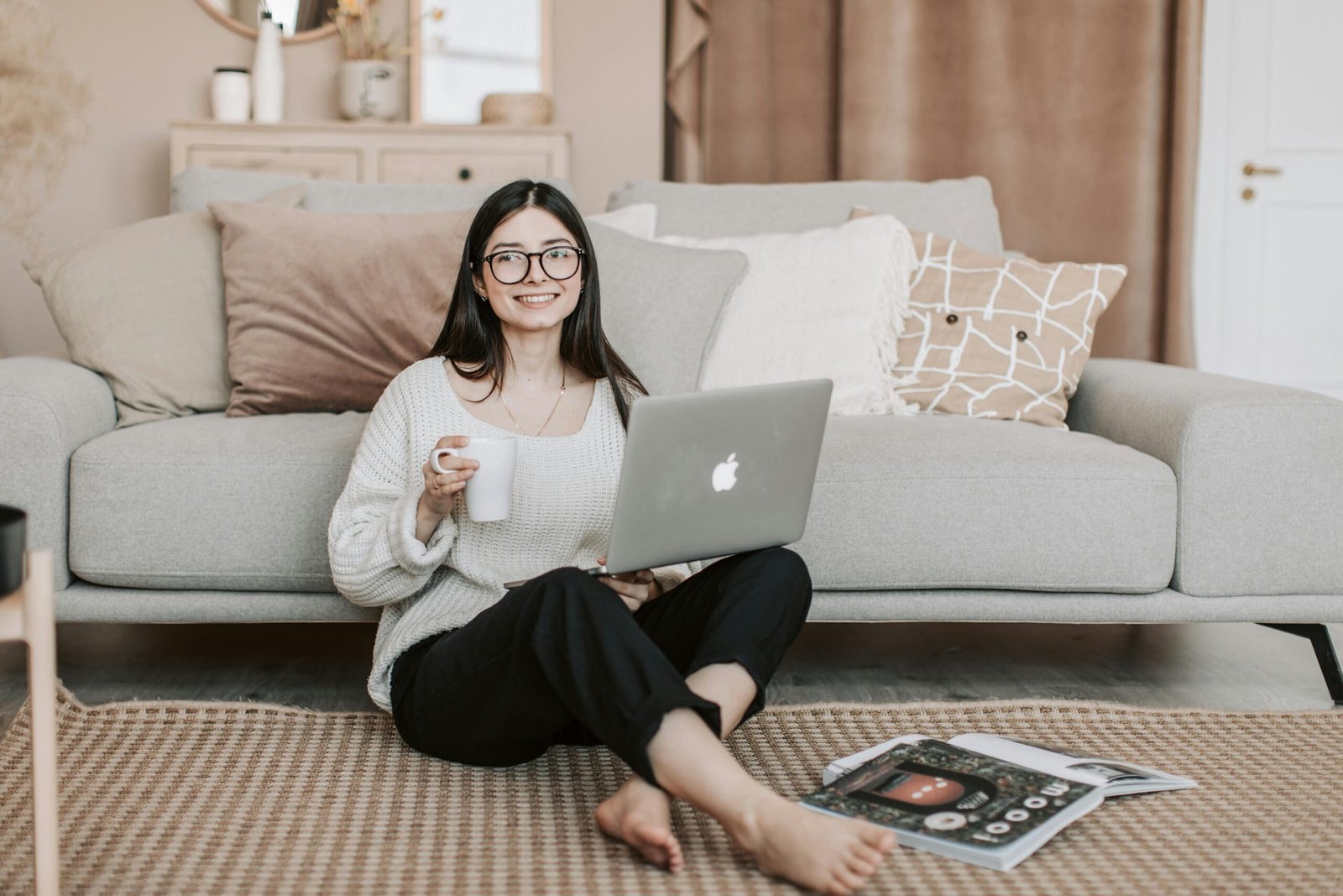 Happy woman enjoying coffee while using a laptop in a cozy living room setting.